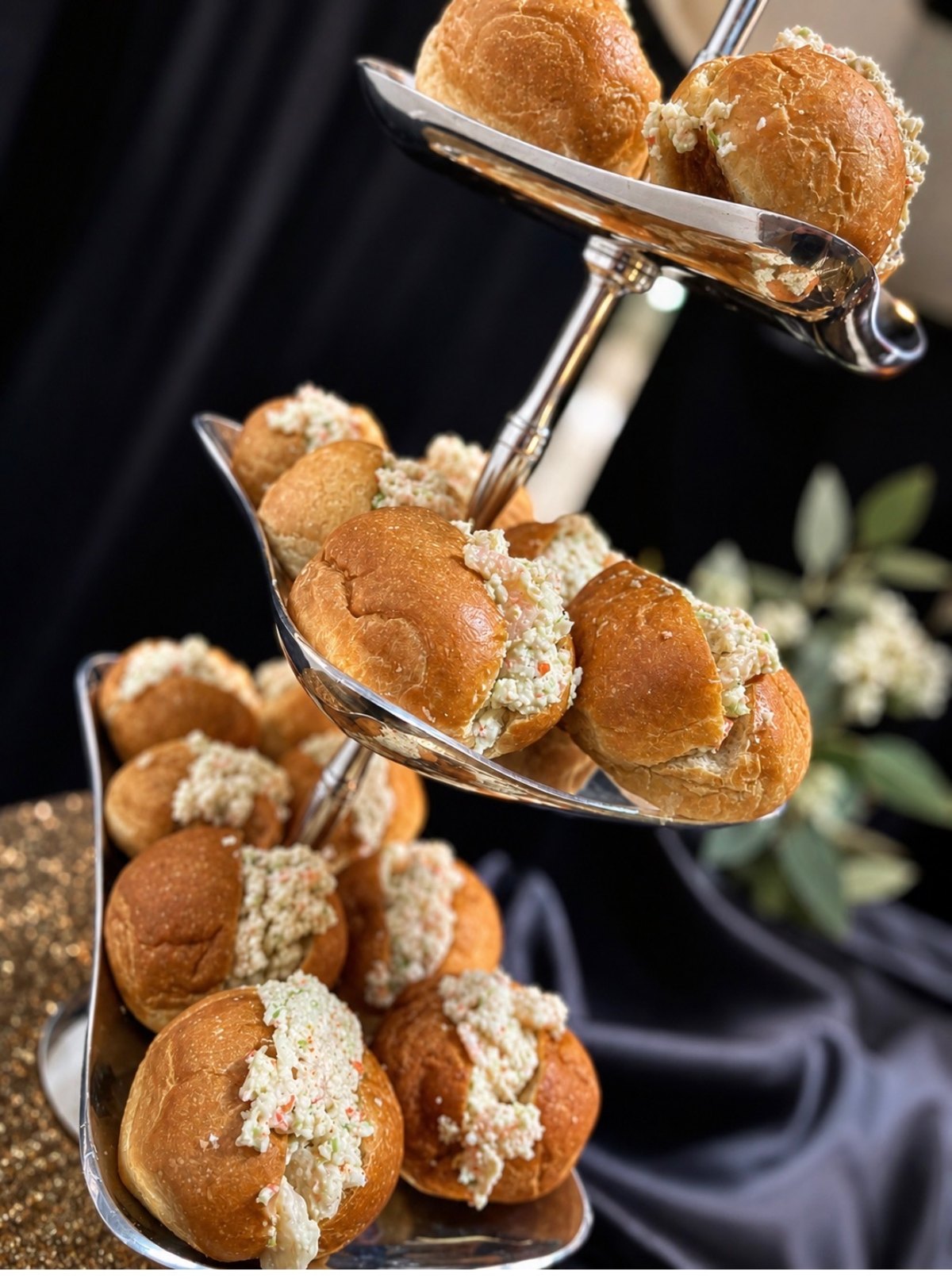 Silver tiered stand of brioche slider rolls filled with seafood salad, styled with olive branches against black and gold linens