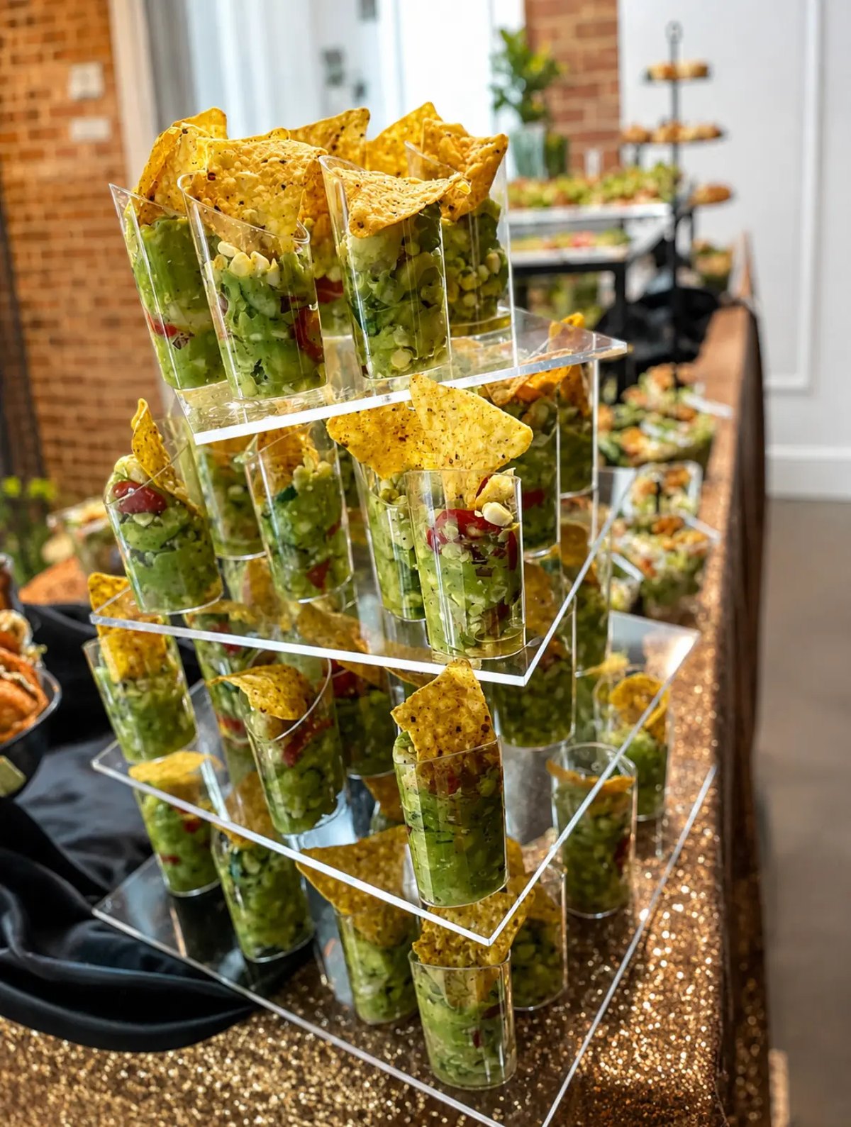 Tiered acrylic display of individual guacamole and corn salsa cups with seasoned tortilla chips on a gold sequin linen at a Sugar & Sparkles catered event