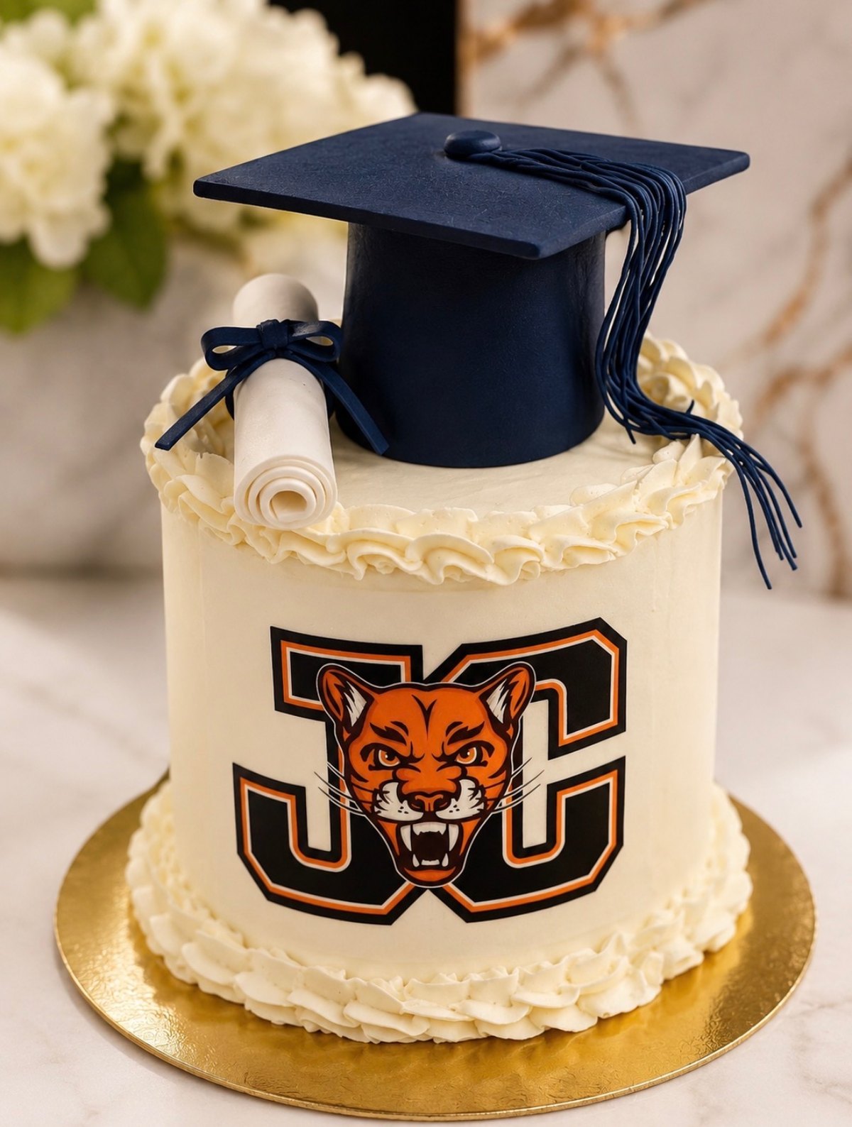 Custom graduation cake in ivory buttercream with a navy fondant cap, sugar diploma scroll and edible school mascot crest on a gold board