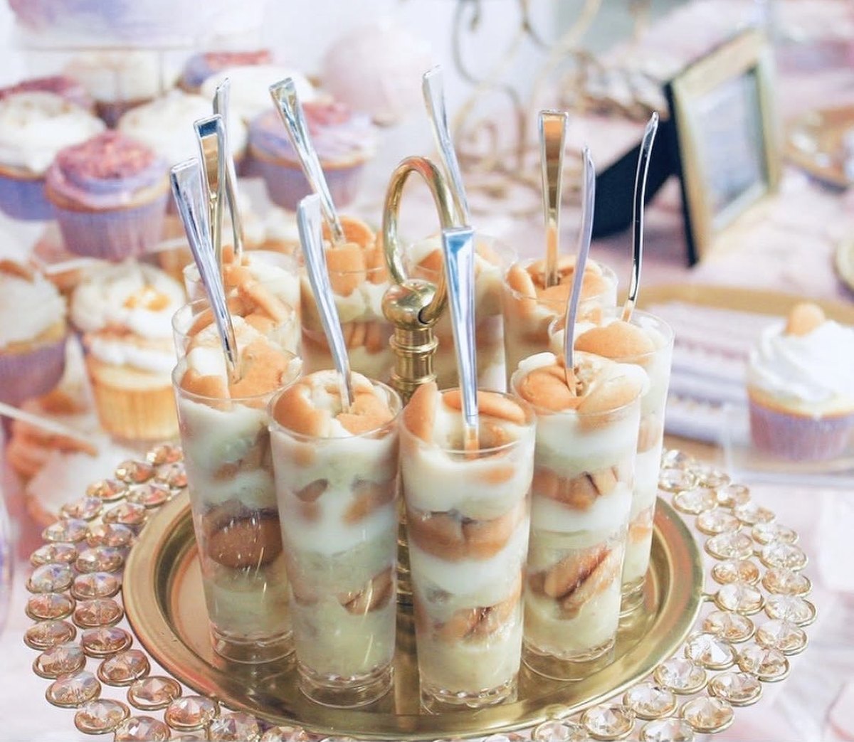 Layered banana pudding shooters with vanilla wafers on a gold and crystal tray beside assorted cupcakes at a dessert table
