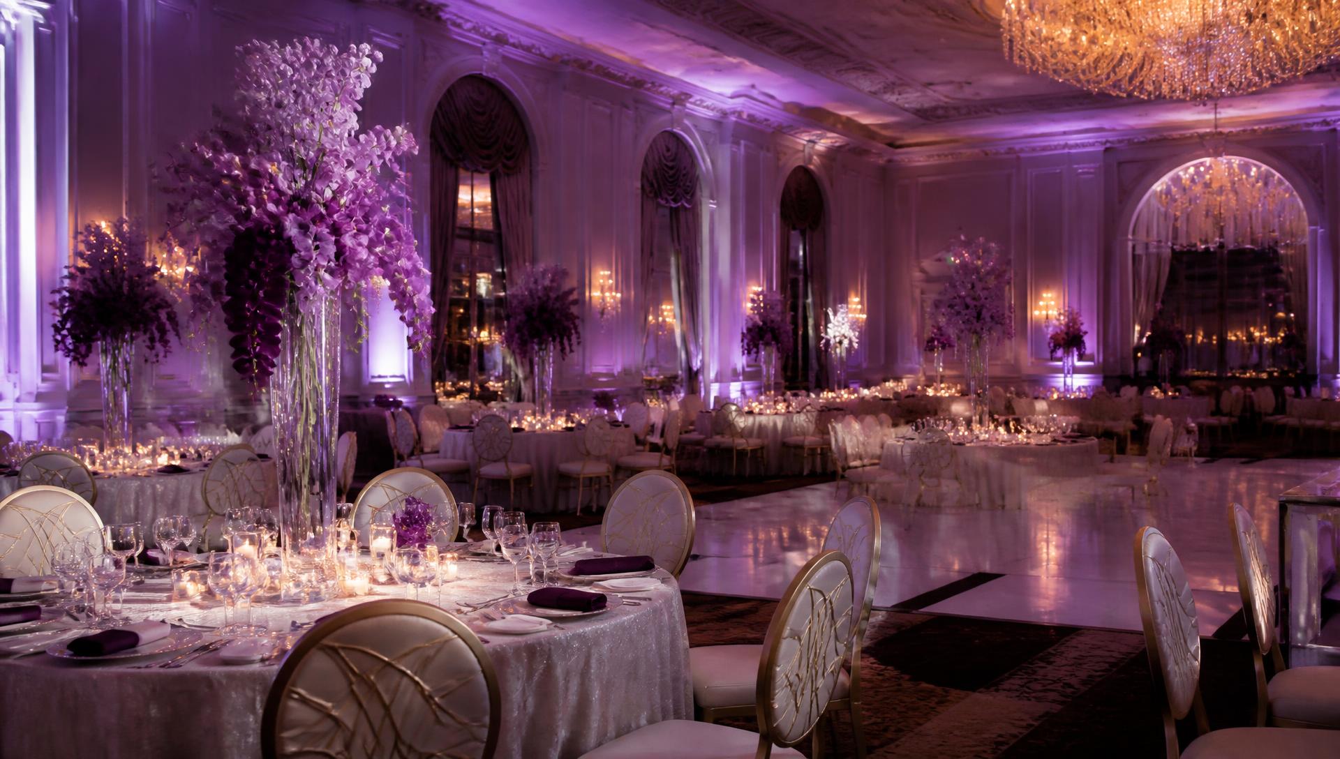 Candlelit luxury ballroom reception with tall lavender orchid centerpieces, violet uplighting and crystal chandeliers
